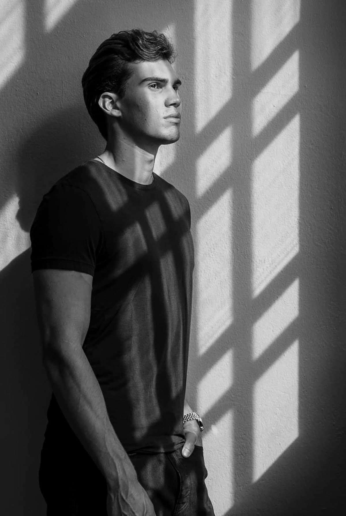 Aleksi Hartikainen standing against a bare wall, lit by hard diagonal bars of light falling through a window blind.
