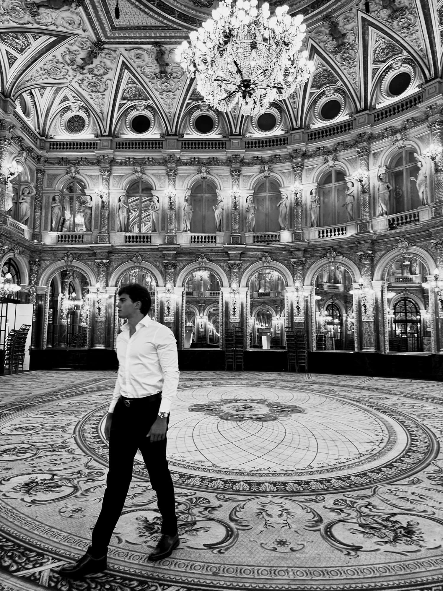 Aleksi Hartikainen crossing the floor of an empty nineteenth-century ballroom, beneath a chandelier and a tier of statues.